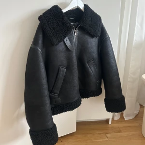 Svart skinnjacka med pälsdetaljer - Snygg svart skinnjacka med mjuka pälsdetaljer på krage, ärmslut och nederkant. Jackan har en dragkedja framtill och två fickor. Perfekt för kyligare dagar med sin stilrena design och varma material.