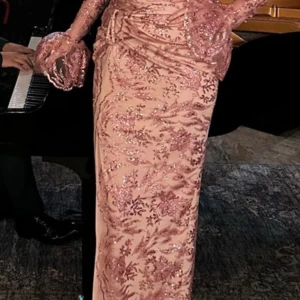 Rosa glittrig långklänning - Elegant rosa långklänning med glittriga detaljer och blommigt mönster. Klänningen har långa ärmar och en slits framtill. Perfekt för en glamorös kväll och studentbalen. 