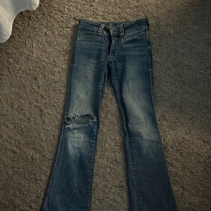 Blå bootcut jeans från H&M - Snygga blå bootcut jeans från H&M med slitningar på ena benet. De har en klassisk femficksdesign och stängs med dragkedja och knappar. Perfekta för en avslappnad stil.