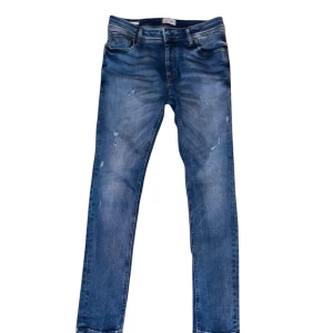 Blåa jack&jones jeans med slitningar  - Snygga blå jeans med en lätt slitning för en trendig look. De har en klassisk femficksdesign och en dragkedja framtill. Perfekta för en avslappnad stil.