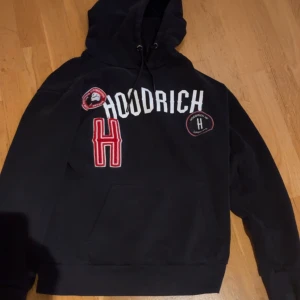 Svart hoodie från Hoodrich - Säljer en svart hoodie från Hoodrich med vita och röda detaljer. Den har en stor H-logga och text på framsidan samt en praktisk ficka. Perfekt för en avslappnad stil.