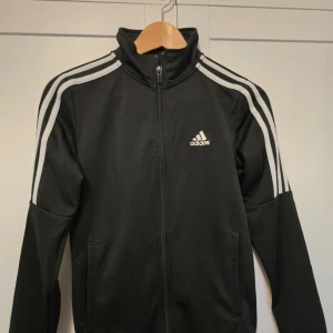 Svart träningsjacka från Adidas - Säljer en svart träningsjacka från Adidas med de klassiska tre ränderna längs ärmarna. Jackan har dragkedja framtill och en hög krage. Passar för dam och herr 