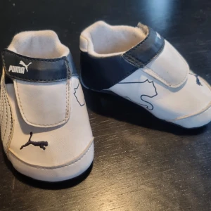 Vita och blå babyskor från Puma - Söta babyskor från Puma i vitt och blått med kardborreband för enkel på- och avtagning. Skorna har Pumas ikoniska logga på sidan, bak, fram och sulan, vilket ger en sportig look. Perfekta för de minsta fötterna.