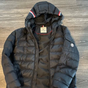 Svart dunjacka från Moncler - Säljer en snygg svart dunjacka från Moncler med huva och dragkedja. Jackan har en cool detalj med röd och vit rand på huvan. Perfekt för kyliga dagar! Köpt här men den var för stor varför den får gå vidare, jättefin! 