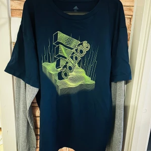 Mörkblå tröja från Adidas - Säljer en mörkblå tröja från Adidas med ett coolt grafiskt tryck i neonfärger på framsidan. Tröjan  har en loose fit i färger mörkblå, grön och grå, perfekt för en avslappnad stil. Tillverkad i mjukt material för extra komfort.