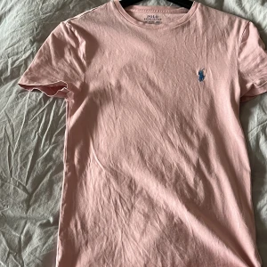 Rosa t-shirt från Polo Ralph Lauren - Säljer en snygg rosa t-shirt från Polo Ralph Lauren med en liten blå logga på bröstet. T-shirten är i en custom slim fit och är tillverkad i Turkiet. Perfekt för en stilren och avslappnad look.