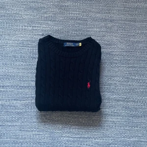Svart kabel stickad tröja  Ralph Lauren (Aldrig använd) - Säljer en stilren svart stickad tröja från Ralph Lauren med kabelstickat mönster. Tröjan har en klassisk rund hals och en liten röd logga på bröstet. Perfekt för kyligare dagar!