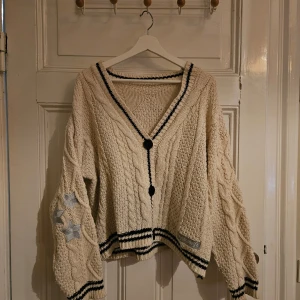Taylor Swift FOLKLORE kofta  - Säljer en mysig beige stickad Folklore kofta. Koftan har svarta knappar och detaljer vid ärmslut och nederkant. Den är dekorerad med stjärnor på ärmarna och har en v-ringad design. Perfekt för kyliga dagar! Obs inte från Taylors Officiella hemsida. Oversized fit.