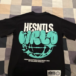 Svart t-shirt från HESNTLS - Snygg svart t-shirt från HESNTLS med stort tryck i turkos och vitt på ryggen. Framsidan har ett mindre tryck med samma färger. Perfekt för en avslappnad stil.