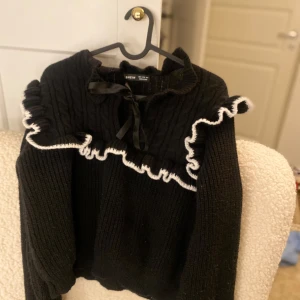 Svart stickad tröja med volangdetaljer - Säljer en svart stickad tröja från Shein med vita volangdetaljer runt halsen. Tröjan har långa ärmar och en söt rosett framtill. Perfekt för kyliga dagar.