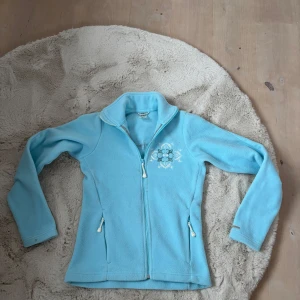 Fleece - Säljer en ljusblå skid fleecejacka från Helly Hansen med broderade detaljer på bröstet och ryggen. Jackan har en dragkedja framtill och två fickor med dragkedjor. Lite fläckar men annars bra.
