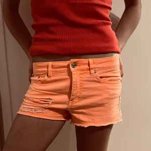 Orange jeansshorts - Snygga orange jeansshorts med slitna detaljer på ena benet. Perfekta för en somrig look. De har en knapp och dragkedja framtill samt klassiska fem fickor.