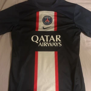Svart fotbollströja från Nike PSG - Säljer en svart fotbollströja från Nike med korta ärmar. Tröjan har ett rött och vitt vertikalt mönster på framsidan och logotypen för Paris Saint-Germain samt Qatar Airways tryckt på bröstet. Perfekt för fotbollsfans som vill visa sitt stöd för laget.