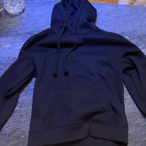 Syna world hoodie - Säljer en svart hoodie med ett coolt tryck på huvan. Tröjan har en klassisk design med dragsko i huvan och en stor ficka framtill. Perfekt för en avslappnad stil.