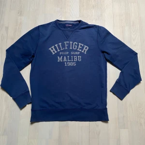 Blå sweatshirt från Hilfiger - Säljer en snygg blå sweatshirt från Hilfiger med texten 'Hilfiger Prep Surf Malibu 1985' tryckt på framsidan. Tröjan har långa ärmar och en klassisk rund halsringning. Perfekt för en avslappnad stil. Strl XL