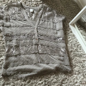 Grå spetsblus från Vila - Säljer en elegant grå spetsblus från Vila med v-ringning och knappar framtill. Blusen har ett vackert mönster och är dekorerad med spetsdetaljer. Perfekt för en stilfull look.