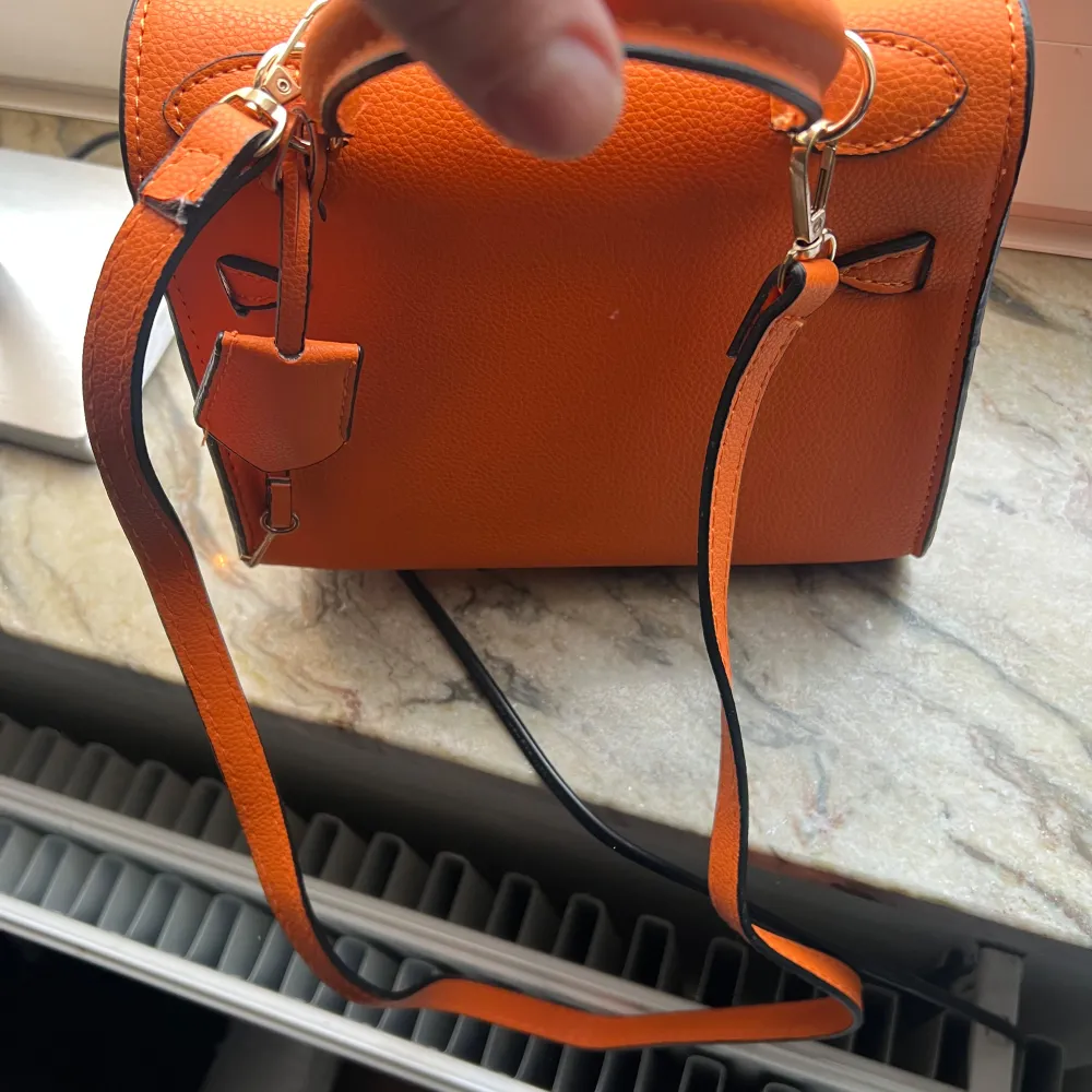 Snygg orange axelväska med en unik låsdetalj framtill. Väskan har en justerbar axelrem och ett handtag för olika bäralternativ. Tillverkad i ett stilrent material med metallbeslag som ger en lyxig känsla. Perfekt för att ge en färgklick till din outfit.. Laukut & Käsilaukut.