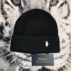 Svart ribbad mössa från Polo Ralph Lauren - Snygg svart ribbad mössa från Polo Ralph Lauren med broderad logotyp i vitt. Perfekt För ger en stilren look. Märkesetikett medföljer. Fram logan har liten defekt på staven 