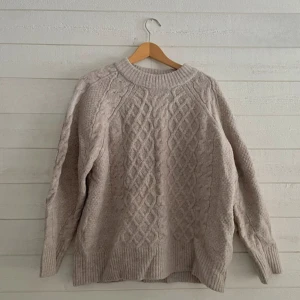Beige stickad tröja - Mysig beige stickad tröja med flätmönster. Perfekt för kyliga dagar med sin varma och bekväma design. Tröjan har långa ärmar och ribbstickade muddar vid ärmslut och nederkant.