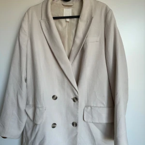 Beige kavaj med knappar - Snygg beige kavaj med dubbelknäppning och klassisk krage. Den har långa ärmar och två fickor framtill. Perfekt för en stilren look. Storlek M