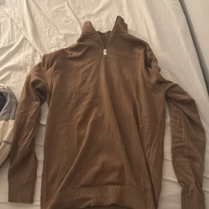 Brun tröja med dragkedja från Jack & Jones - Säljer en brun tröja från Jack & Jones med dragkedja vid halsen. Tröjan har långa ärmar och ribbade muddar vid ärmslut och nederkant. Perfekt för en stilren och bekväm look.