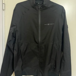 Svart jacka från Sail Racing - Snygg svart jacka från Sail Racing med dragkedja framtill. Jackan har en stilren design med långärmade ärmar och en diskret logga på bröstet. Perfekt för en sportig look.