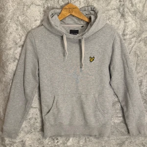 Grå hoodie från Lyle & Scott - Säljer en stilren grå hoodie från Lyle & Scott med deras ikoniska logga på bröstet. Tröjan har en klassisk känguruficka och justerbar huva med snören. Perfekt för en avslappnad stil.