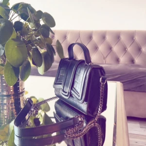 Svart axelväska med kedjedetaljer - Snygg svart axelväska i skinn med quiltad design och kedjedetaljer. Perfekt för att ge din outfit en edgy touch. Väskan har ett praktiskt handtag och en justerbar axelrem för bekväm användning.