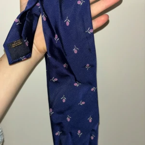 Blå slips med blommigt mönster från Eton - Snygg blå slips från Eton med ett diskret blommigt mönster i rosa. Tillverkad i Italien av 100% siden, vilket ger en lyxig känsla och fin glans. Perfekt för att ge en elegant touch till din outfit.