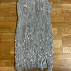 Silver glittrig klänning från NLY One - Snygg silverfärgad klänning från NLY One med glittrande paljetter och en elegant slits. Axelbandslös design som passar perfekt för festliga tillfällen. Klänningen har en dragkedja baktill för enkel påklädning.