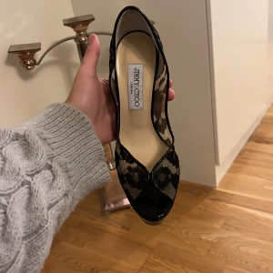 Svarta spetsiga pumps från Jimmy Choo - Eleganta svarta pumps från Jimmy Choo med spetsdetaljer och hög klack. Tillverkade i Italien, dessa skor har en sofistikerad design med öppen tå och en smal siluett.