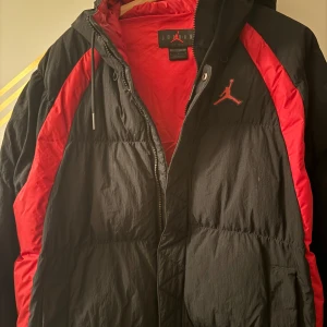 Svart och röd puffer jacka Jordan -   Jackan har en dragkedja framtill och en huva med dragsko. Perfekt för kyliga dagar. Jackan är skön och håller dig varm. finns inte mycket kvar av denna jacka.  Helst upphämtning. Storlek M-L