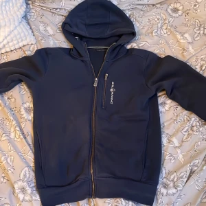 Mörkblå hoodie från Sail Racing - Säljer en mörkblå hoodie från Sail Racing med dragkedja och huva. Perfekt för kyliga dagar med sin mjuka insida. Den har en liten logga på bröstet och en praktisk ficka med dragkedja.