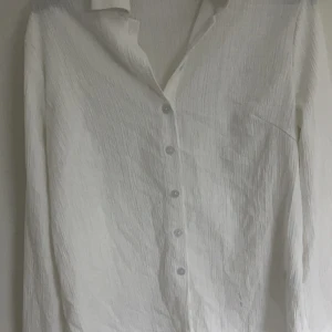 Vit långärmad skjorta - Säljer en stilren vit långärmad skjorta med knappar. Skjortan har en krage och ett subtilt texturerat mönster som ger en elegant touch. Perfekt för en klassisk look. Den är genomskinlig därför säljer jag den. 