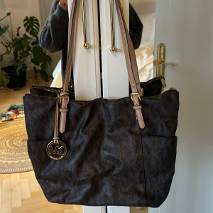 Michael Kors väska  - Snygg brun axelväska från Michael Kors med beige axelremmar och ett dekorativt MK-hänge. Väskan har en praktisk dragkedja upptill och är perfekt för att bära dina dagliga nödvändigheter.Väskan har en fläck på insidan, därav det billiga priset!