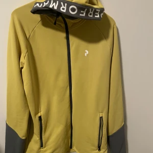 Peak Performance träningströja- gul/grå - Snygg gul hoodie med grå detaljer och dragkedja framtill. Perfekt för en sportig look med text på huvan. Långärmad design med praktiska fickor. Använd fåtal gånger därmed mycket fint skick, Storlek : S