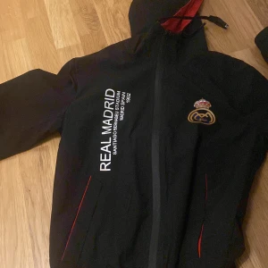 Svart Real Madrid jacka med huva - Snygg svart jacka med Real Madrid-tryck och emblem. Jackan har en huva med röd insida och dragkedja framtill. Perfekt för fotbollsfans!      Nypris:1049