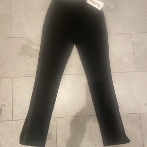 Svarta byxor från Hope, modell Move - Snygga svarta byxor från Hope i modellen 'Move Trousers'. De har en elegant och stilren design med hög midja och raka ben med revärer samt en 20 slits vid ankeln. Perfekta för en chic och modern look. Inneben längd 80 cm. Längd från midjan 110 cm. Runt midjan i navelhöjd 38 cm