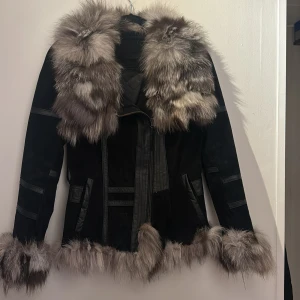 Svart jacka med pälsdetaljer - Snygg svart jacka med lyxiga pälsdetaljer på krage, ärmslut och nederkant. Jackan har en figursydd passform och stängs med dragkedja. Perfekt för att hålla sig varm och stilren under kyliga dagar. Den är i mocka och läder. Pälsen är räv. 