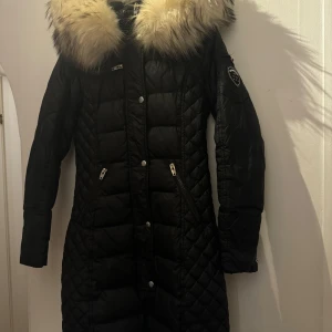 Svart quiltad dunjacka med pälsdetalj - Snygg svart quiltad dunjacka med stor pälsdetalj på kragen. Jackan har dragkedja och knappar framtill samt långa ärmar. Perfekt för kalla vinterdagar. Rock and blue med vit tvättbjörns krage. En reva på insidan men går att sy ihop annars är det bra skick! 