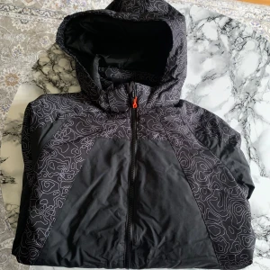 Svart jacka med mönster från H&M - Snygg svart jacka från H&M med ett unikt mönster och luva. Jackan har dragkedja och praktiska fickor framtill. Perfekt för kyligare dagar med sitt mjuka foder.