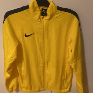 Gul träningsjacka från Nike - Säljer en gul träningsjacka från Nike med Dri-Fit-teknologi. Jackan har en dragkedja framtill och svarta detaljer på axlarna samt en svart Nike-logga på bröstet. Perfekt för sport och träning. Även helt ny