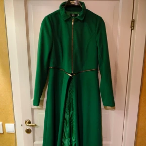 Grön lång kappa med dragkedja - Fantastisk grön lång kappa med dragkedja framtill. Perfekt för höst och vår. Kappan har en elegant design med långa ärmar och en stilren krage. Oanvänd!