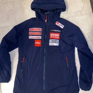 Blå jacka med logotyper - Säljer en blå jacka från Huski Wear med flera logotyper, inklusive Bauhaus, Cramo, Skistar och Audi. Jackan har en dragkedja framtill och en praktisk huva. Köpt från Stadium Outlet. Bara använd några gånger!