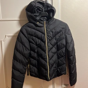 Svart pufferjacka från Svea - Snygg svart pufferjacka från Svea med huva och dragkedja i guld. Jackan har ett quiltat mönster och är perfekt för kyligare dagar. Den har långa ärmar och en liten patch på ärmen.