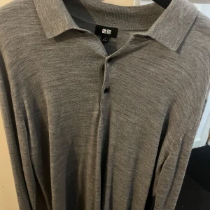Grå ulltröja från Uniqlo - Säljer en stilren grå tröja från Uniqlo i 100% ull. Tröjan har en klassisk krage och knappar framtill, perfekt för en elegant look. Passar bra till både vardag och mer formella tillfällen.