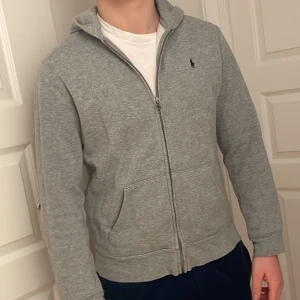 Grå hoodie från Polo Ralph Lauren - Säljer en stilren grå hoodie från Polo Ralph Lauren med dragkedja och broderad logga på bröstet. Perfekt för en avslappnad look. 