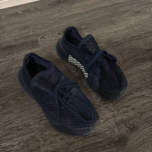 Blåa sneakers från Adidas - Säljer ett par svarta sneakers från Adidas med ett unikt nätmönster och snörning. Skorna har en sportig design med en bekväm passform, perfekt för vardagsbruk.