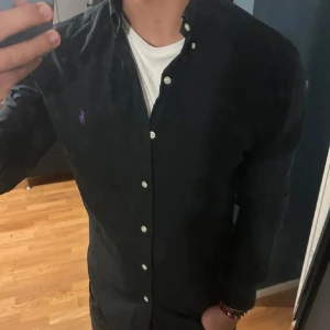 Ralph Lauren Skjorta - Snygg svart skjorta från Ralph Lauren med lila broderad logga på bröstet. Skjortan har långa ärmar och knappar framtill. Perfekt för en stilren look.