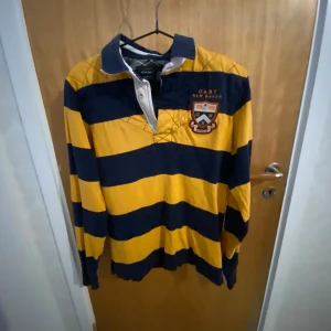 Randig rugbytröja från GANT - Säljer en klassisk randig rugbytröja från GANT i blått och gult. Tröjan har långa ärmar och en broderad logga på bröstet. Perfekt för en sportig look med en quiltad detalj på baksidan.